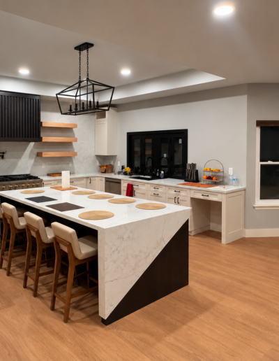 Finished open-concept kitchen remodel featuring a large waterfall island, white shaker cabinets, floating wood shelves and custom black range hood