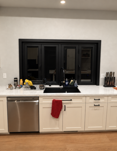 Finished kitchen sink area with white shaker cabinetry, stone-look countertops, black-framed window and stainless steel dishwasher